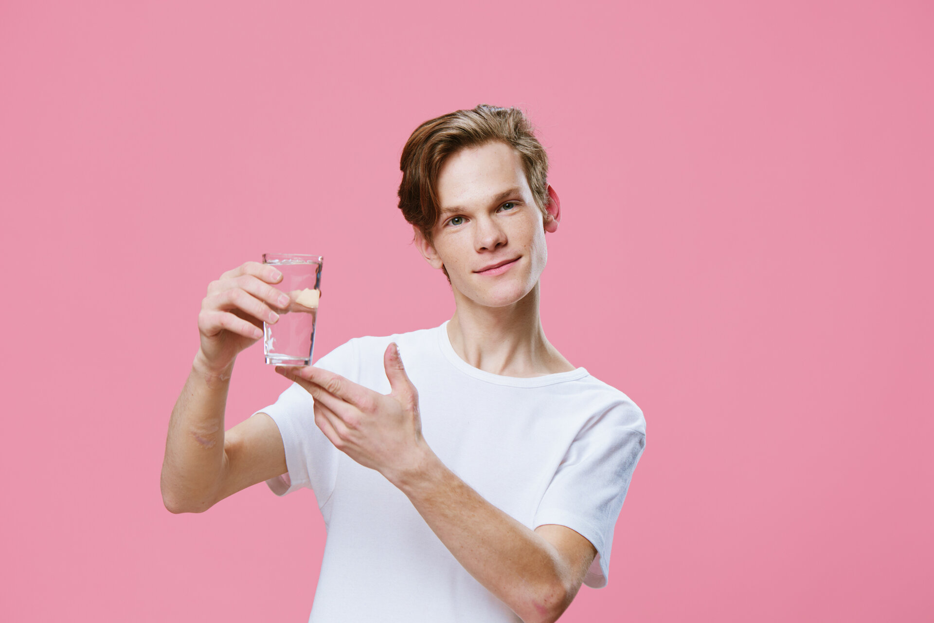 Ein sympathischer Typ in einem weißen T-Shirt zeigt auf ein Glas Wasser in seiner Hand.