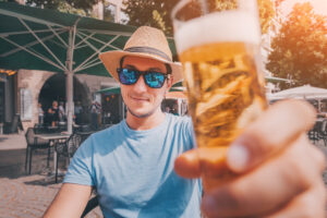 Glücklicher Tourist mit Sonnenbrille und Hut, der traditionelles kölsches Bier in einer Kneipe im Freien probiert und trinkt